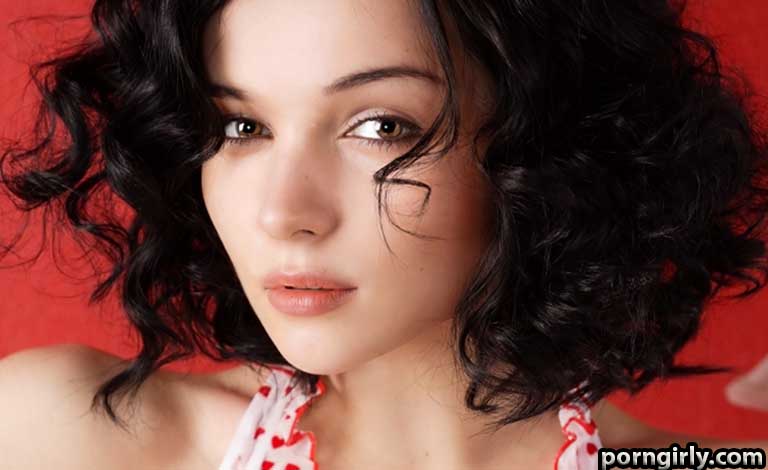 Close-up portrait of a young woman with dark curly hair against a red backdrop, looking at the camera.