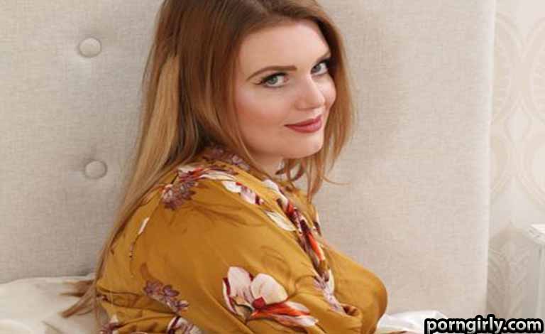 Woman with long blonde hair sits on a bed, wearing a mustard floral robe and smiling at the camera, turned slightly over her shoulder.